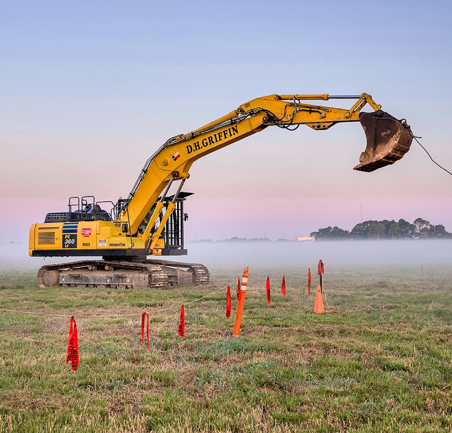 Yellow excavator working on a grassy field at sunrise.